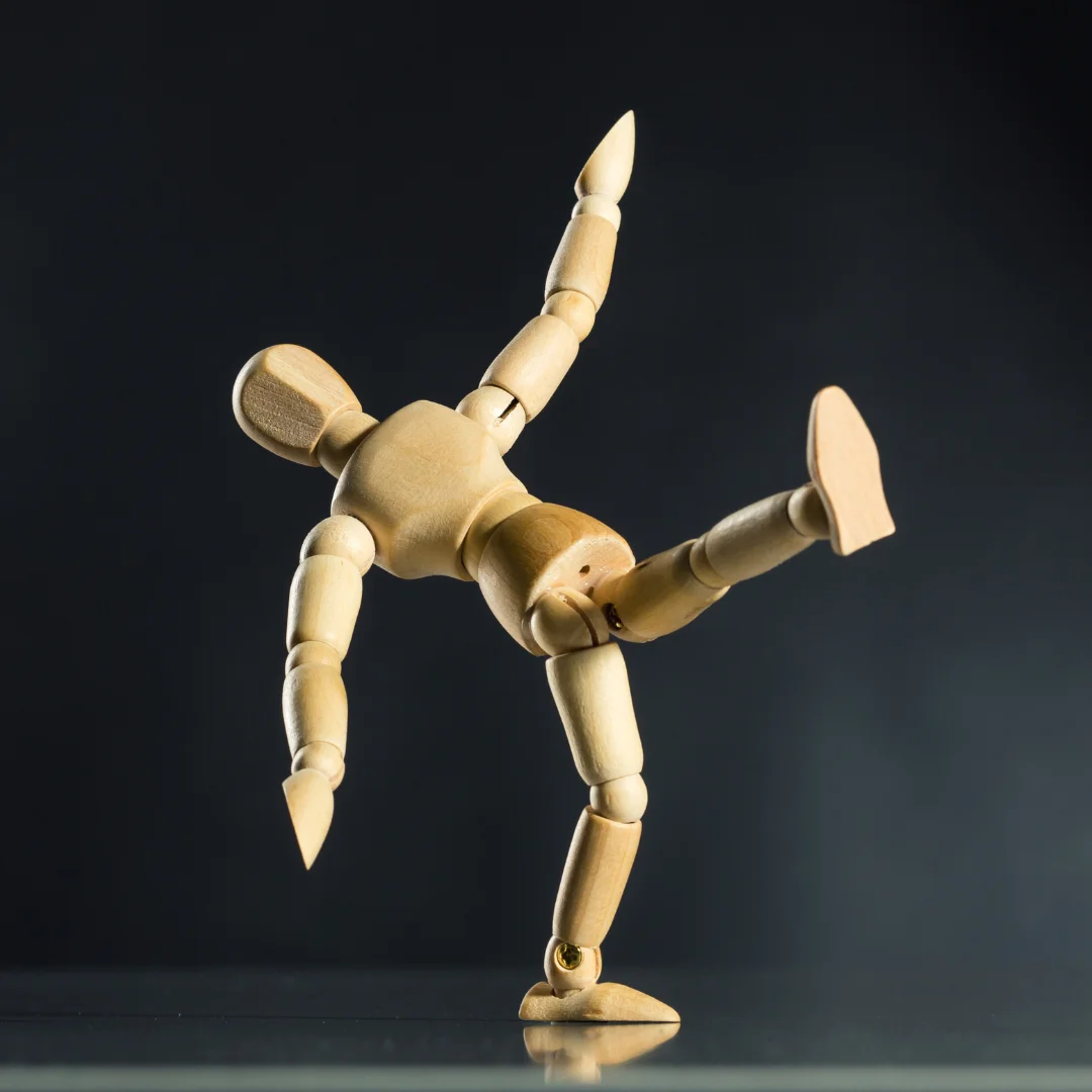 A single Manikin is standing in the center of the frame with one leg up like he is balancing. His arms are spread out to balance himself. There is a black smokey background and a drop shadow below him