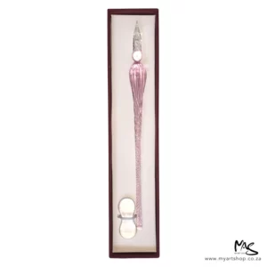 There is a single Glass Dipping Pen Pink in a box, shown vertically, in the center of the frame. The box is brown, and there is a beige cushioning in the bottom of the box and the pen is sat on the cushioning. There is a clear glass pen rest, placed at the bottom of the pen. The image is on a white background.