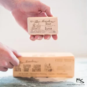 A persons hands are seen coming in from the left hand side of the frame. Their top hand is holding a sealed bar of Masters Hand Soap. There is a large block of something in the background, but the image is blurred. The image is cut off by the frame.