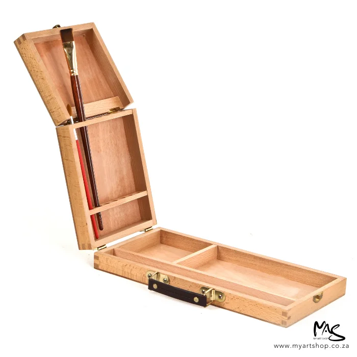 An open Wooden Paint Brush Box is shown in the center of the frame. The hinged lid is facing the left hand side of the frame and the base is along the bottom of the frame. There is a single brush being displayed in the box, the brush is held in the lid of the box. the box is wooden and is on a white background