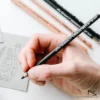 A persons hand is seen coming in from the right hand side of the frame holding a General's Carbon Sketch Pencil. There are 4 pencils in the background, behind the hand that are blurred, and thee is a piece of paper with a sketched drawing also behind the hand, along the left hand side of the frame, it is also blurred. The image is cut off by the frame and on a white background.