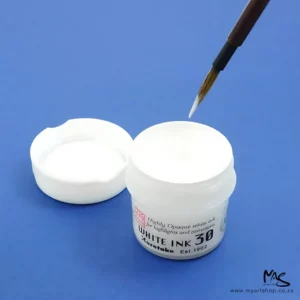 A close up of an open bottle of ZIG Cartoonist White Ink with a paint brush being dipped into the ink. The lid is to the back left of the bottle. The bottle is white and the lid is white and the ink is white. On a blue background.
