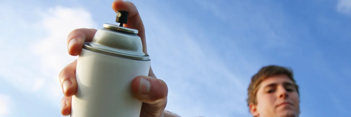 Stick it Right: Find the Perfect Spray Adhesive for Your Fabric Masterpieces just like this guy is doing in the image. there is a man to the right of the image you can see his face and he is looking towards the camera, he has short blond hair. you can see his hand is holding a can. the top of the can is visable with his finger on the nozzle of the can about to push it. the can is white and silver with a white nozzle. the background in the image is a blue sky