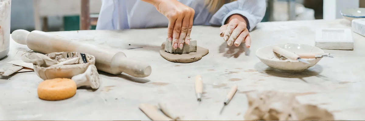 Clay Creativity Unleashed Unconventional Air Dry Clay Ideas is what is being shown in this image. there are a pair of hands in the center of the image cutting shapes into the clay using various tools. her hands are covered in dry cly and next to her are various tool srtewn about the table infront and around where she is working. she is wearing a light blue long sleeve shirt. on the table is a long brown rolling pin and a bowl with two tools resting in it. the table is a white ish colour and the background is a blurred visual of the room.