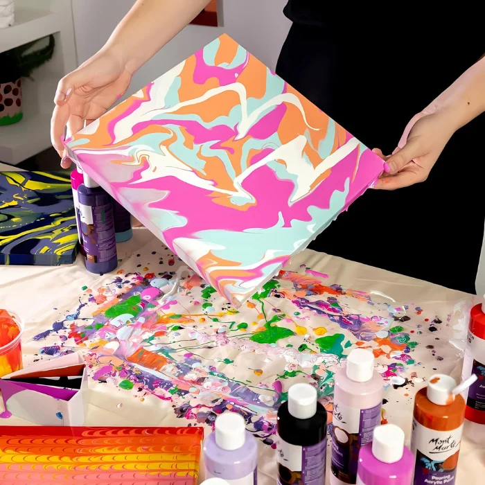 mont-marte-pouring-acrylic-paint-07 A persons hands are seen coming in from the top of the frame holding a canvas. They are moving it to make the paint run. There are a few colours of paint on the canvas and drips underneath the canvas. There are various other bottles of pouring paint on the table below the canvas.