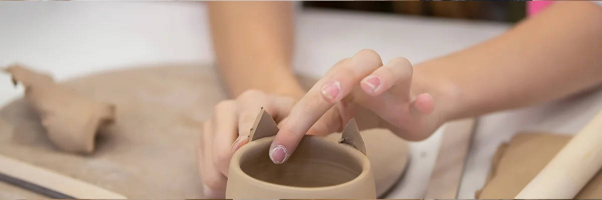 Sculpting Sensation Unveiling the Secrets of Air Dry Clay Modelling is demonstrated in this image by the hands of a young artist as she molds and shapes a vase in the centre of the image. the clay is a terracotta colour and you can see her hands molding the clay. there are ears on the other side of the clay. and in a blurred vision behind her you can see her arms and more flat clay that she is working with. in the center of the image on a white background