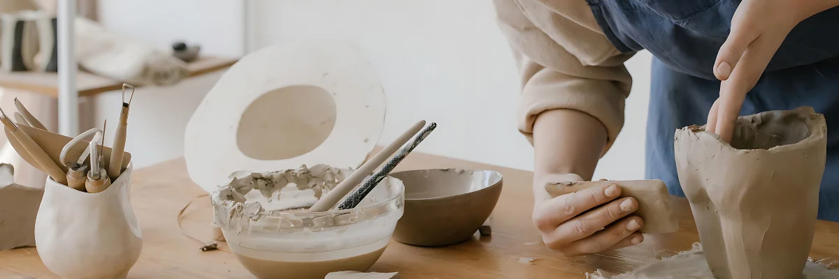 Sculpting Without Limits The Wonders of Air Dry Clay Sculpture is what this lady is doing. there is a lady on the right hand side of the image. you can only see her arms and hands as she works her clay figure that is in front of her. to the left of her in the image are various bits of clay and other clay tools on a big wooden table with a large bowl of dirty clay water in the center of the table. the background is a slightly blurred image of the room she is in.