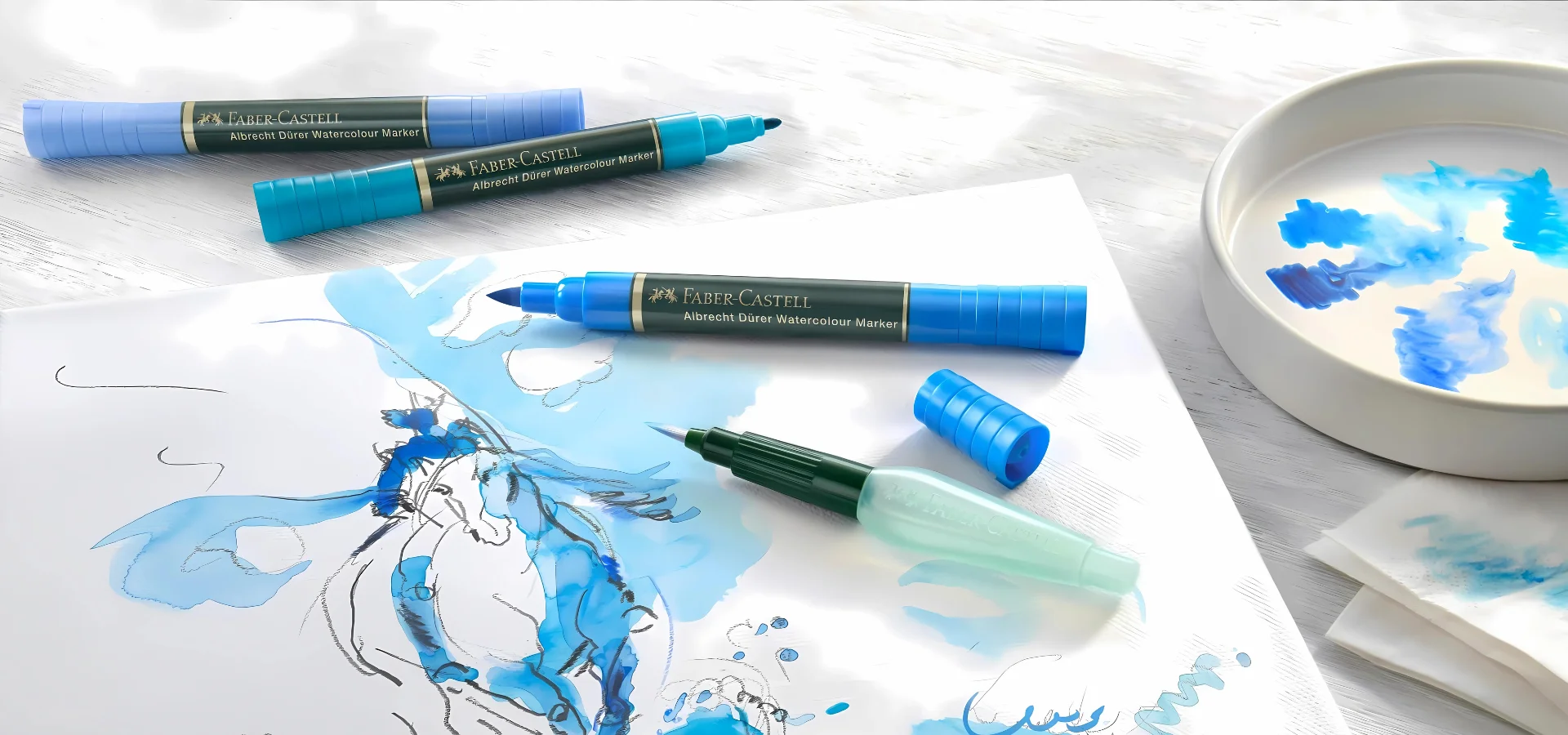 albrecht-durer-watercolour-markers-discover-the-magic/ in this image there is a watercolour canvas in the center and there are a few blue shades of the pens lying on top of it. they have their lids off and are scattered around the canvas. you can see the different nibs on the pens. thye canvas has a drawing on it. the pens are stocked at my art shop