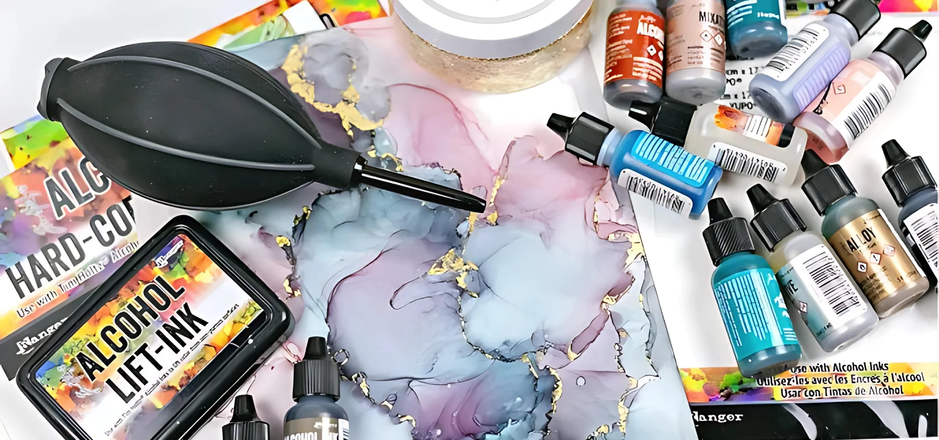 alcohol-ink-accessories-helping-to-conquering-art-challenges shows in the image on the right hand side various alcohol ink bottles that have been used and are placed haphazardly on the yupo paper. in the middle is a art work breated from the alcohol inks in shades of blue and purple with gold leafing effects on it in gold. to the left of that is the alcohol ink blower that is black and bulbus and has a nozzle that is facing towards the bottom right corner of the image. to the left of that is an alcohol lift ink pad that is black with yellow and green label. sitting ontop of yupo paper. behind them and half in the image is a tub of gold leaf.