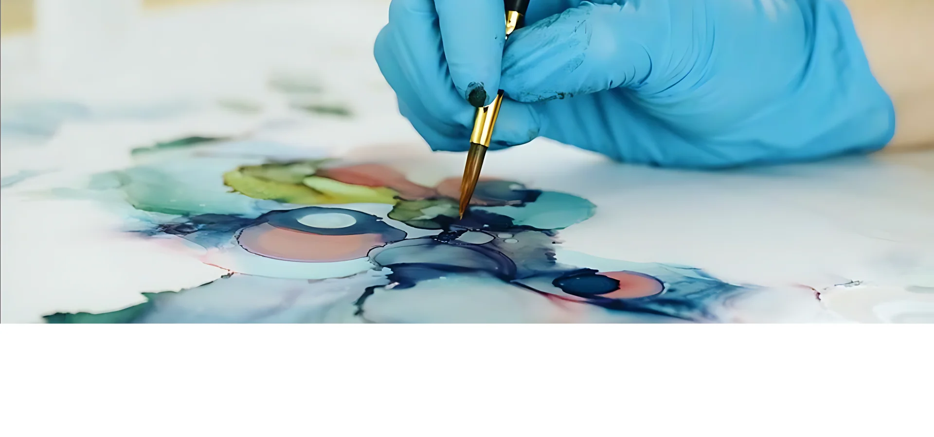 alcohol-ink-surfaces-ink-delights. in this image there is a hand in a blue latex glove coming in from the right hand side of the image. holding a paintbrush and painting on the yupo paper with the alcohol inks. the paper is flat and some of the colours are visable. in blue and greens with a touch of pink. mainly painted in little circles. the background is a blurred white