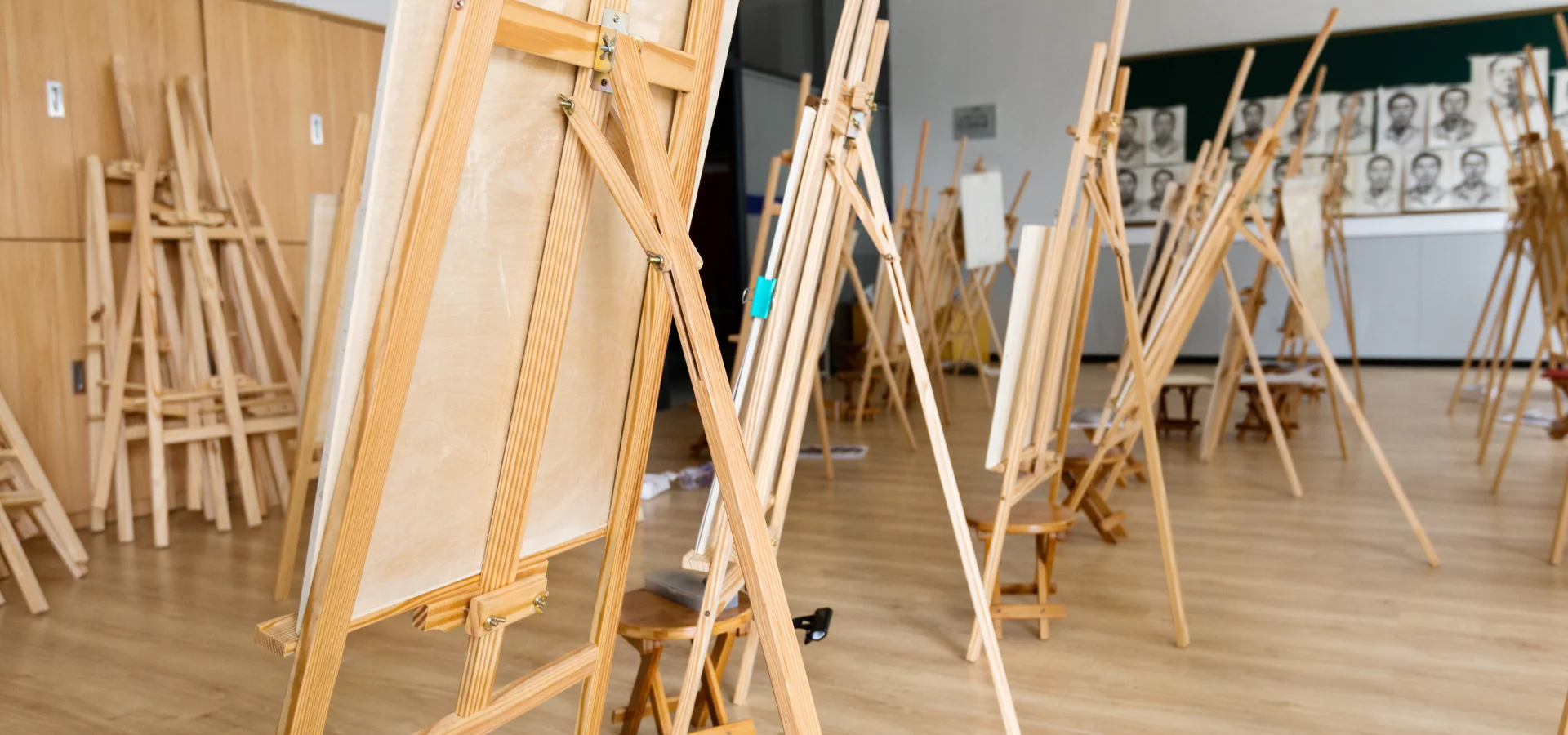 art-easels-for-painting-mastery. in this image there are a selection of easels open and inuse with canvas on them in an art class setting. the easels backs are facinf towards the front of the image. there are easels resting against the wall on the left of the image. at the other end of the image is a board with various drawings on it. the floor the easels are standing on is a light wooden floor. the light is coming from the right hands side.