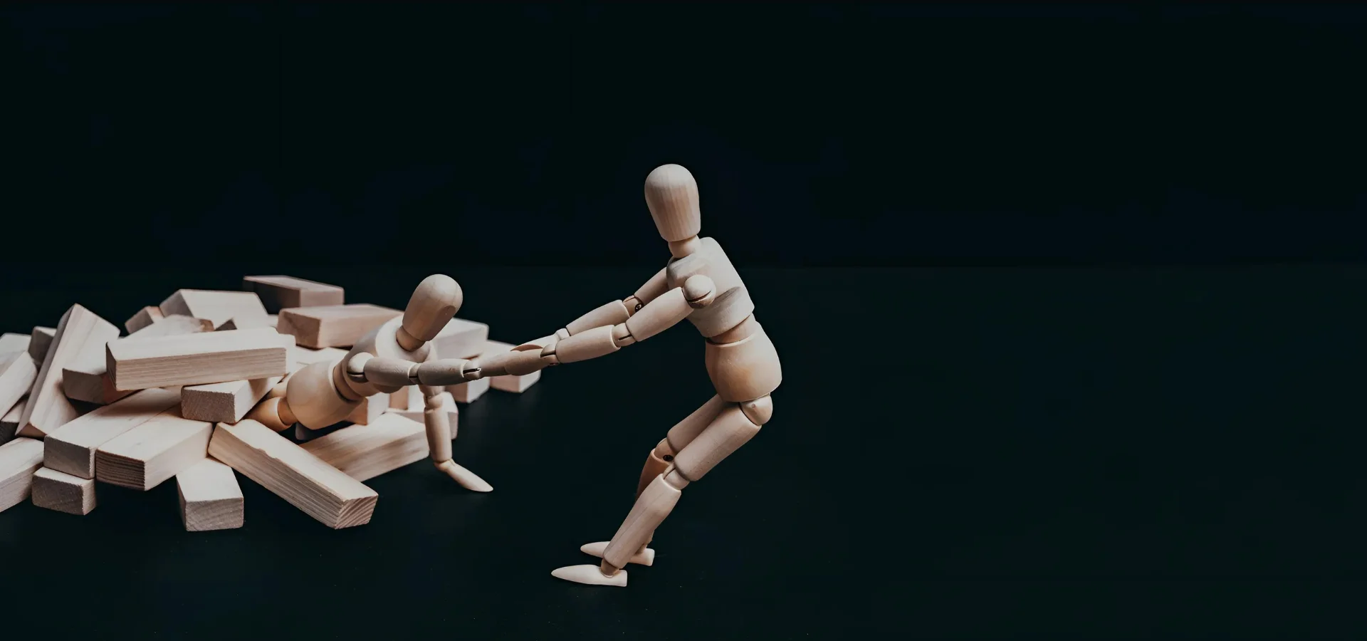 artist-manikins-cracking-the-code. in the centre of the image and to the left hand side there are a pile of little wooden blocks and a wooden manikin is coming out of them with their arm extended as if being pulled out of it. to the right is another manikin with its arms extended as if it is pulling the other one out of the pile of wooden blocks. they are all a light brown wood on a black background. they are sold at my art shop