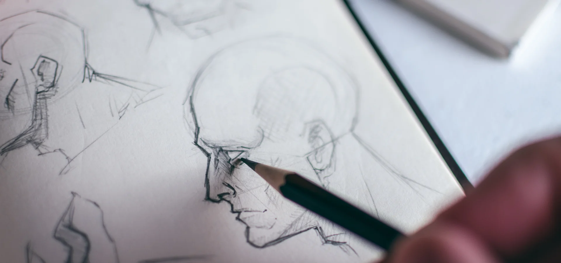 best-sketching-pencils-for-beginner-artists SHOWS A PERSONS HAND IN THE RIGHT BOTTOM CORNER OF THE IMAGE, THEY ARE HOLDIG A BLACK PENCIL AND DRAWING ON PAPER. THEY ARE SKETCHING A PERSONS FACE IN BLACK ON WHITE PAPER. THERE ARE VARIOUS OTHER FACES ON THE PAPER THAT HAVE BEEN DRAWN