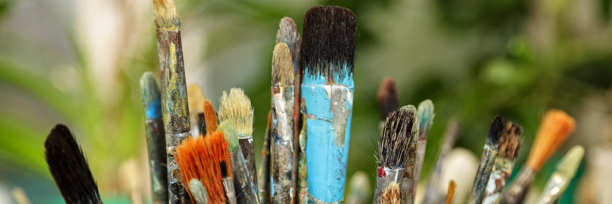 Brushing Off The Mess Paintbrush Cleaning for Quirky Artists shows in the front of the image a variety of very dirty and well used paintbrushes in a bunch of different shapes and sizes. they are all covered in paint on theor brisltes and on their ferrules. they are standing up and you can see them clearly the background is a blurred visual of the garden behind them. the picture is very bright.