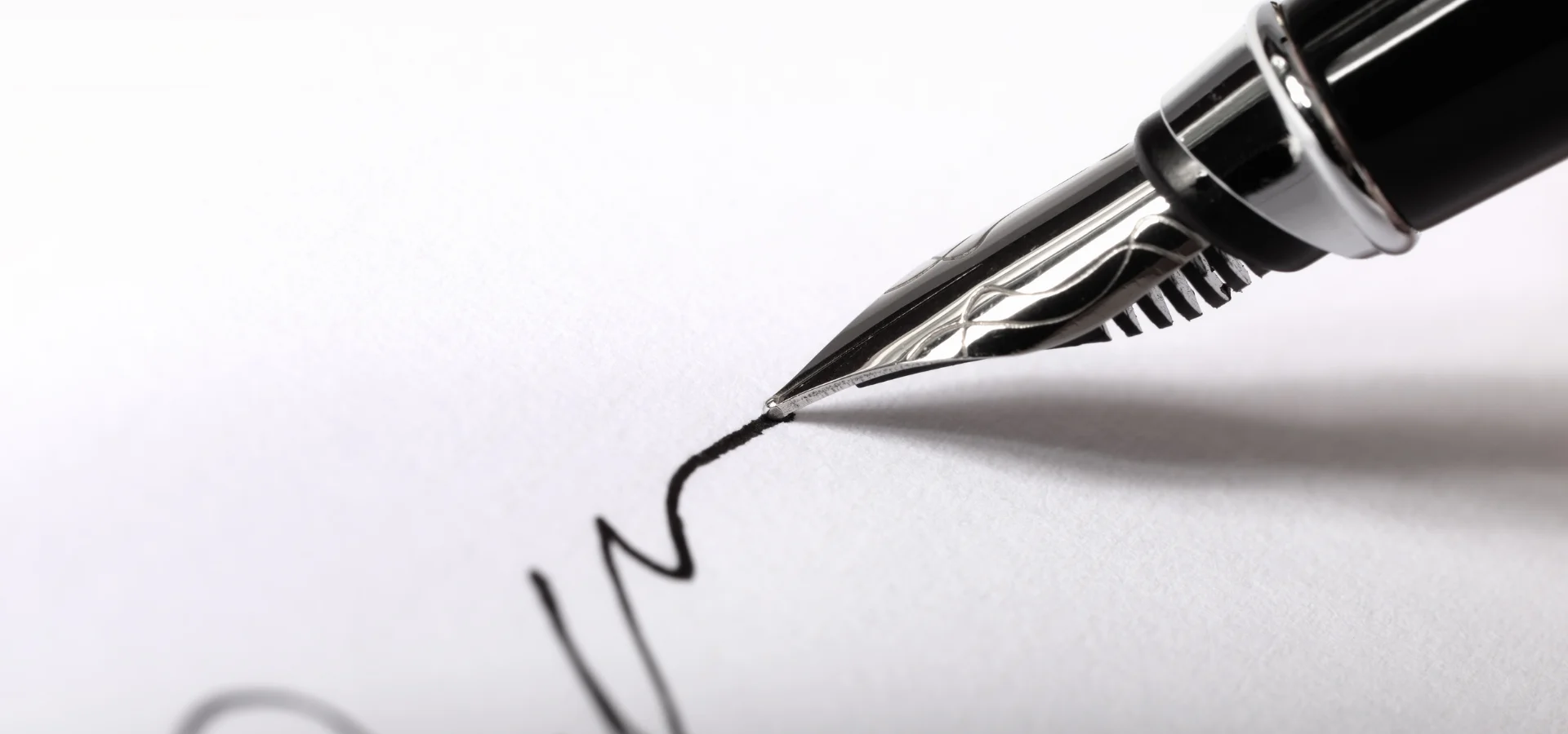 calligraphy-pens-discover-the-magic. in the center of the image is a black pen coming in from the right hand side. it has a silver nib. the pen is touching the paper and black ink is coming from the tip onto the white paper. the pens are sold at my art shop