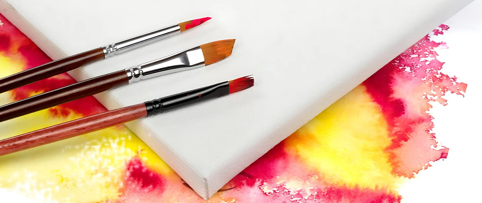 Canvas Quest Hunt for the Ideal Watercolour Canvas is showing to the left of the image three different shaped paintbrushes leaning on a white canvas. the paintbrushes have long red stems and two have silver ferrules one has a black ferrule. they are synthetic watercolour brushes with red paint and orange paint on the bristles. the canvas is uneven in the image. it is resting on a watercolour background of pink and yellow that is bleeding into eachother and blending out to make a large background painting. it is in the center of the image.