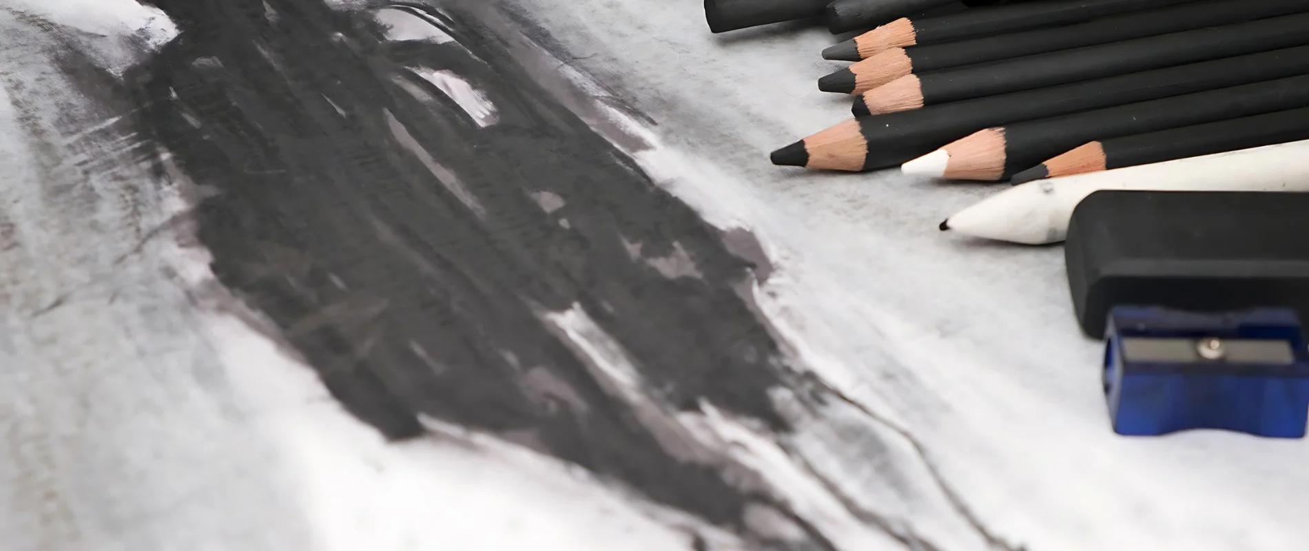 Charcoal and Graphite Drawing Sets A Quest for the Best. in this image at the left hand side you can see a scribble and smudge of black charcoal across the paper. the smudge fades out over to the right where an assortment of charcoal sticks and pencils is laying with the nibs facing the left hand side of the image. the paper is an off white slightly grey colour. there are various tools used for drawing scattered across the page.