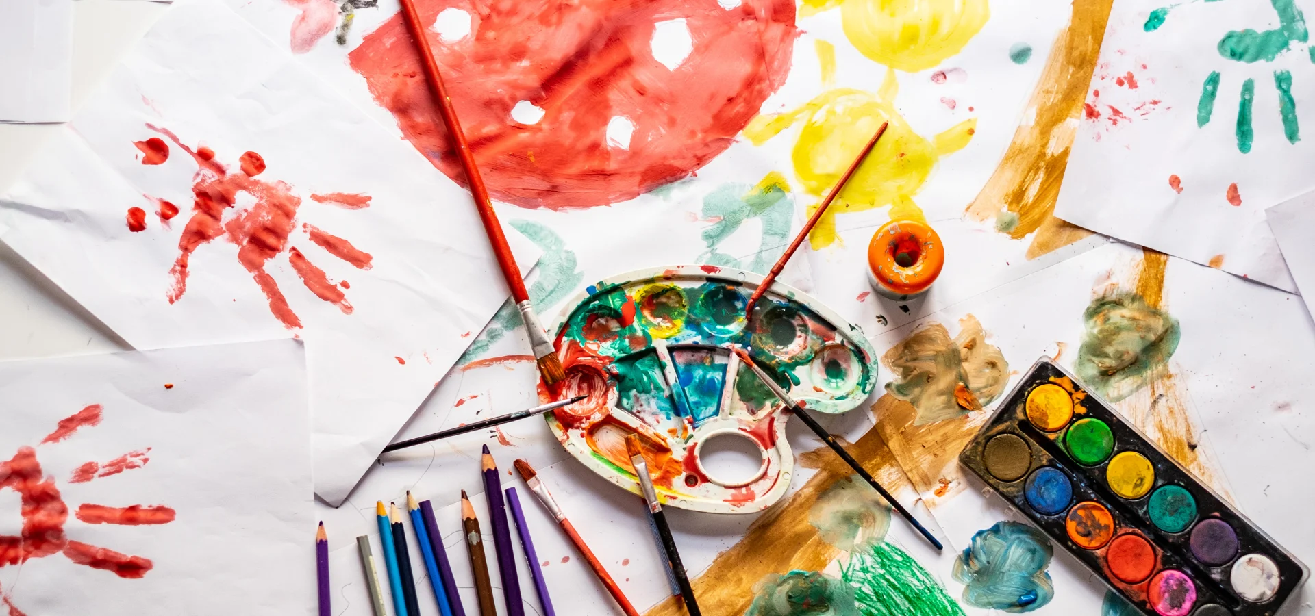 childrens-art-supplies-extravaganza. in this image there are a variety of art supplies on a table. there is a palette that is fulled with paint and has paintbrushes sitting on it, paintings of mushrooms and handprints on the paper in different bright colours. pencils lined up on the left of the palette.