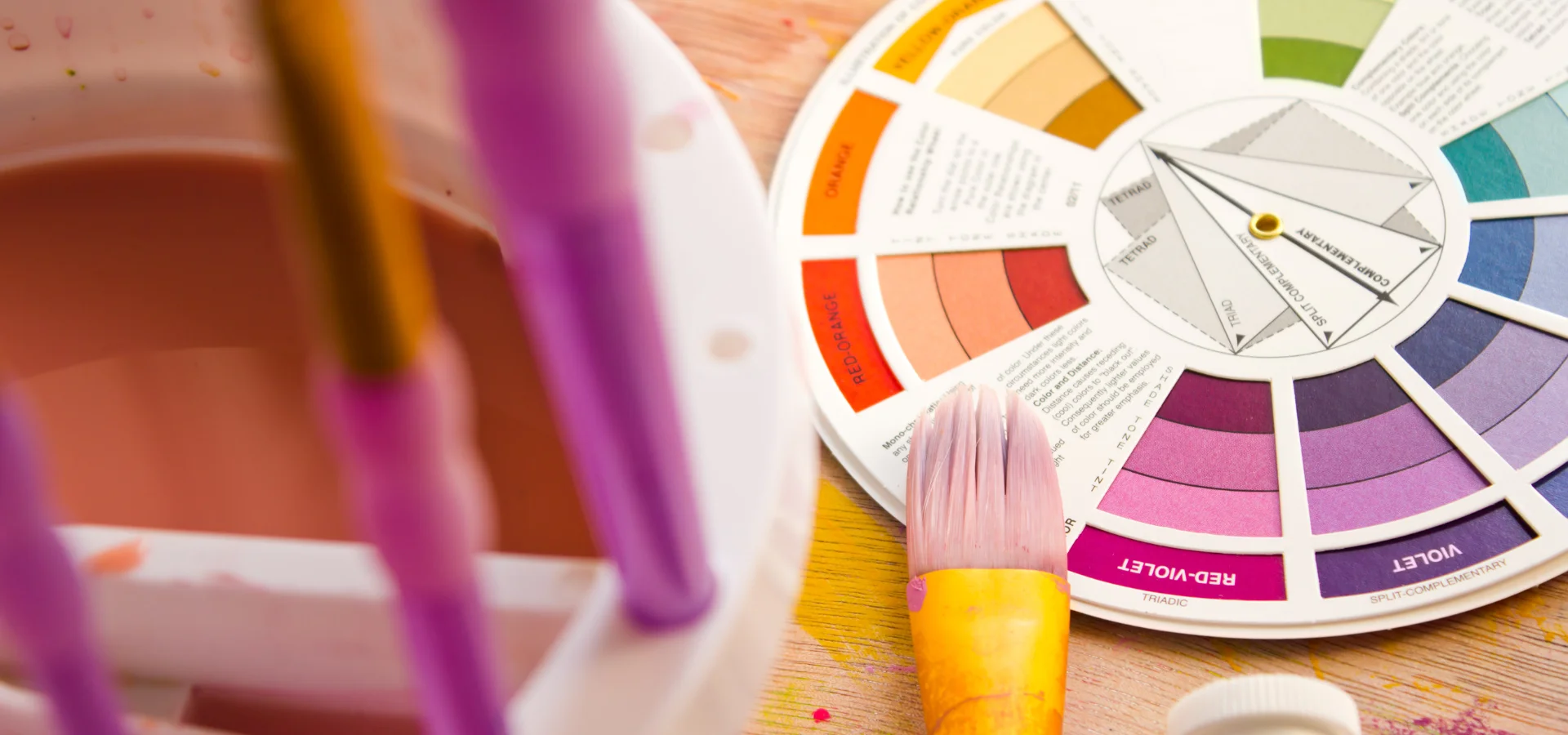 Colour Wheel Chart Antics Unveiled. in this image on the right hand side is a round colour wheel showing the colours that can be mixed. on the left of it is a large flat paintbrush with a yellow ferrule resting on it slightly. behind the brush to the left is the blurred image of a paintbrush holder with other pink stemmed brushes inside of it. they are all lying on a wooden tabletop. the light is coming from the right hand side of the image and casting slight shadows underneath the objects.