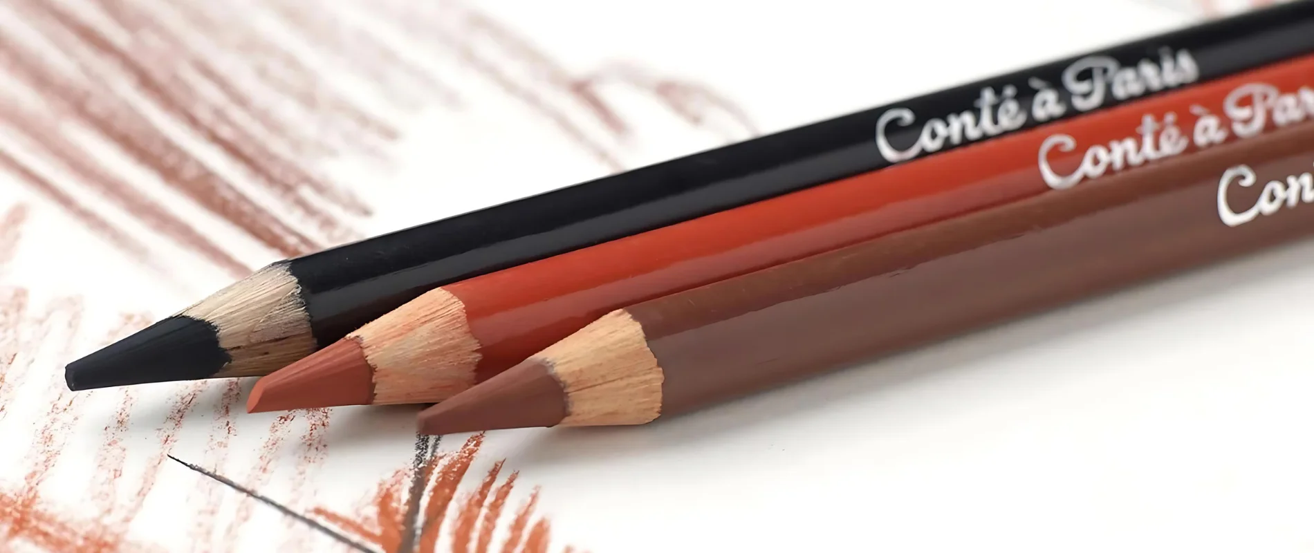 in the center of the image are three colour crayons from conte a paris. they are from left to right. black, red and brown. they ar=ll have sharp nibs that are facing the left bottom corner of the image. they have silver writing visable on them saying the name of the pencil. they are resting on a paer that has been drawn on using those colours.