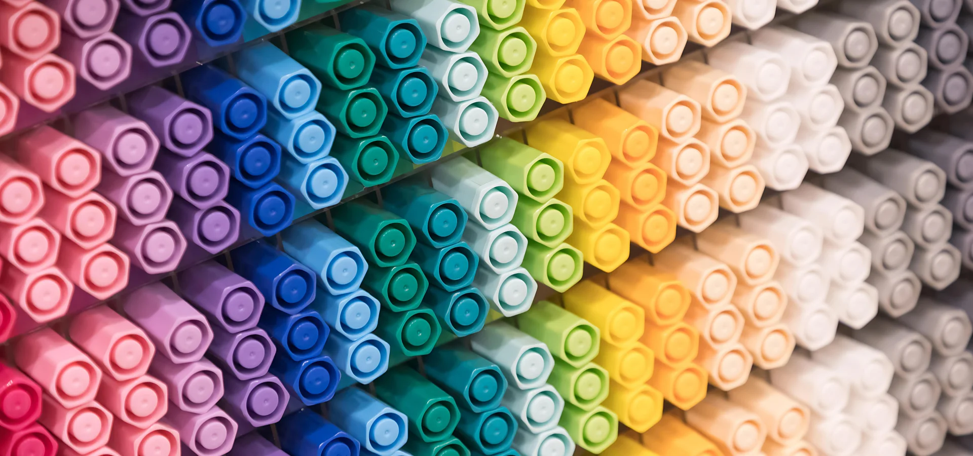 copic-ciao-markers-a-chromatic-delight. across the whole image is a stand filled with many markers in a colour coded fashion. they are in a rainbow order. from pink, purple, blue, green, yellow, cream, white, and grey. at an angle. they take up the whole image. copic ciao markers are sold at my art shop