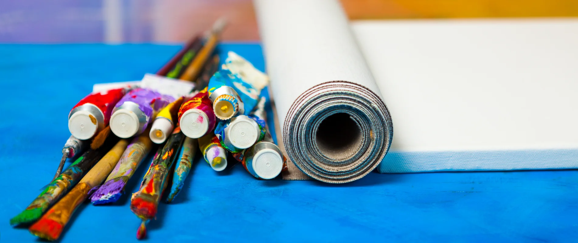 Create Like a Pro Unveiling the Best Canvas Types for You is showing on the right hand side of the image a canvas and next to it a roll of canvas with the top facing the front of the image. next to that on the left hand side are a variety of paint tubes in different colours and paintbrushes covered in paint. they are lying on a blue surdace. the canvas and canvas roll are brilliant white in colour and the the blue surface is a brilliant bright blue. the backgound is a blur of the room behind