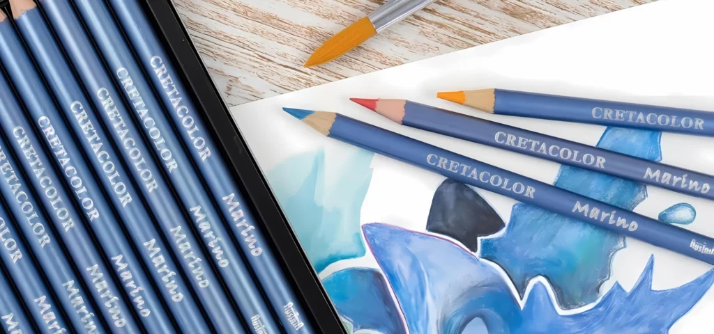 Cretacolor Marino Watercolour Pencil. A close up of a set of pencils coming out of the bottom left hand side of the frame with an image drawn using the pencils along the bottom of the frame and some loose pencils scattered over the drawn picture. And there is a paintbrush above the drawn image. Part of the image is cut off by the frame.