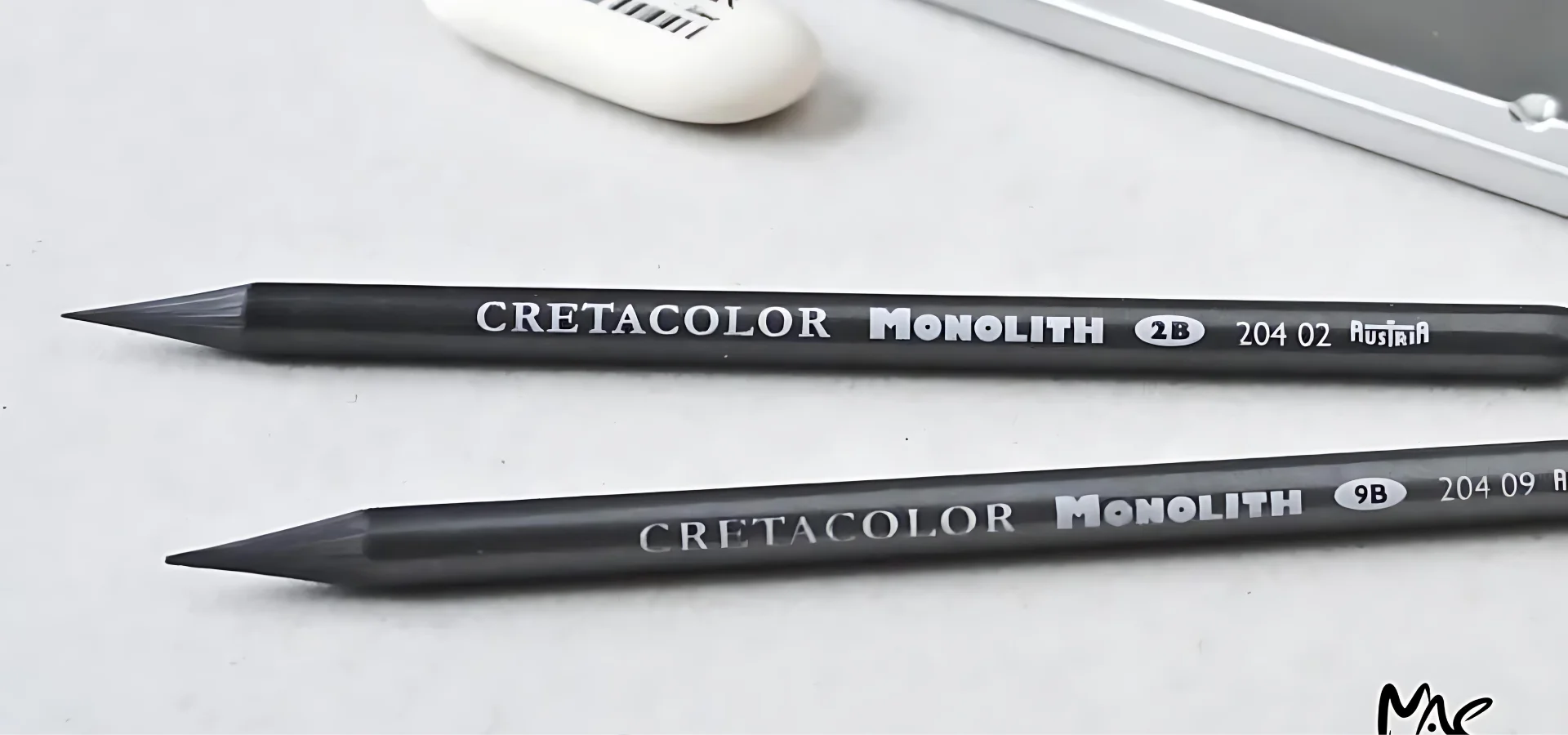 Cretacolor Pencils A Journey shows in the image. two cretacolor monolith graphite pencils that are lying next to eachother at the bottom of the image. they are facing nib point twoards the left. they are s solid grey colour. they have white writing visable on them stating their name and the brand. they are lying infront of a white eraser and a silver metal pencil tin. on a white tabletop.