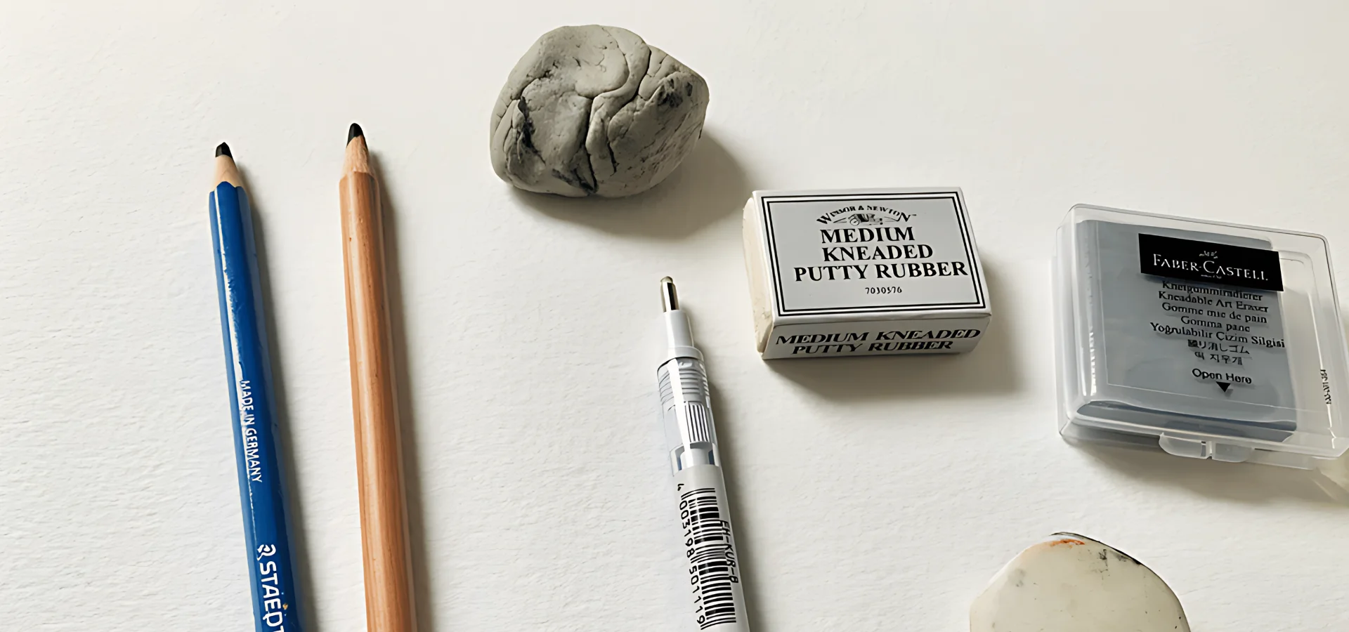 Drawing Accessories Finding the Best for You shows in the image a slection of different types of erasers on a paper. on the left hand side are two different pencils and to the right of it are variuos brands of putty erasers. they are on a white table