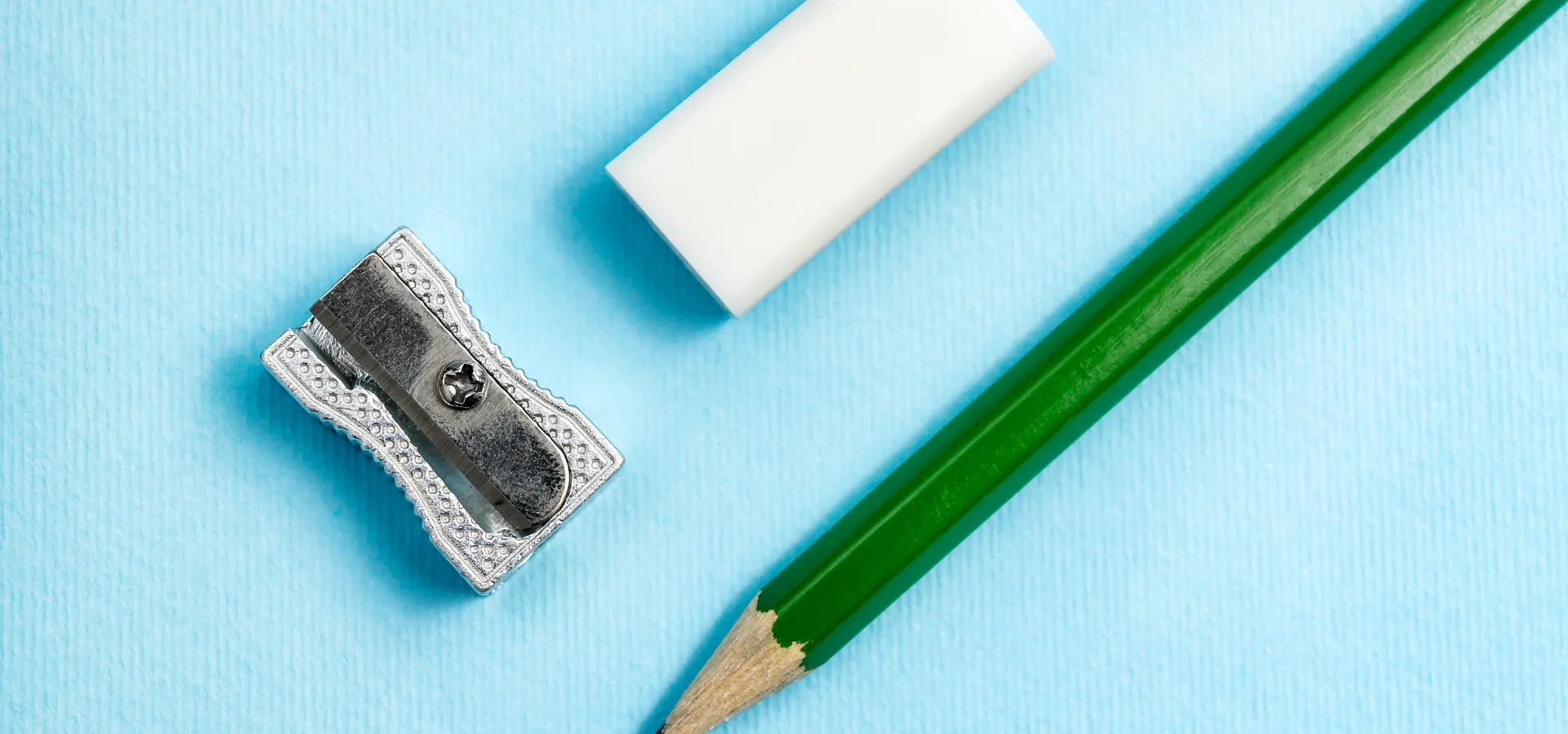 erase-like-a-pro-discover-the-best-erasersshows a pencil sitting at an angle in the center of the image. the pencil has a green casing and a brown wood top with a grey nib. next to it and above it is a rectangular white eraser. infront of that is a single silver sharpener. they are on a light blue background