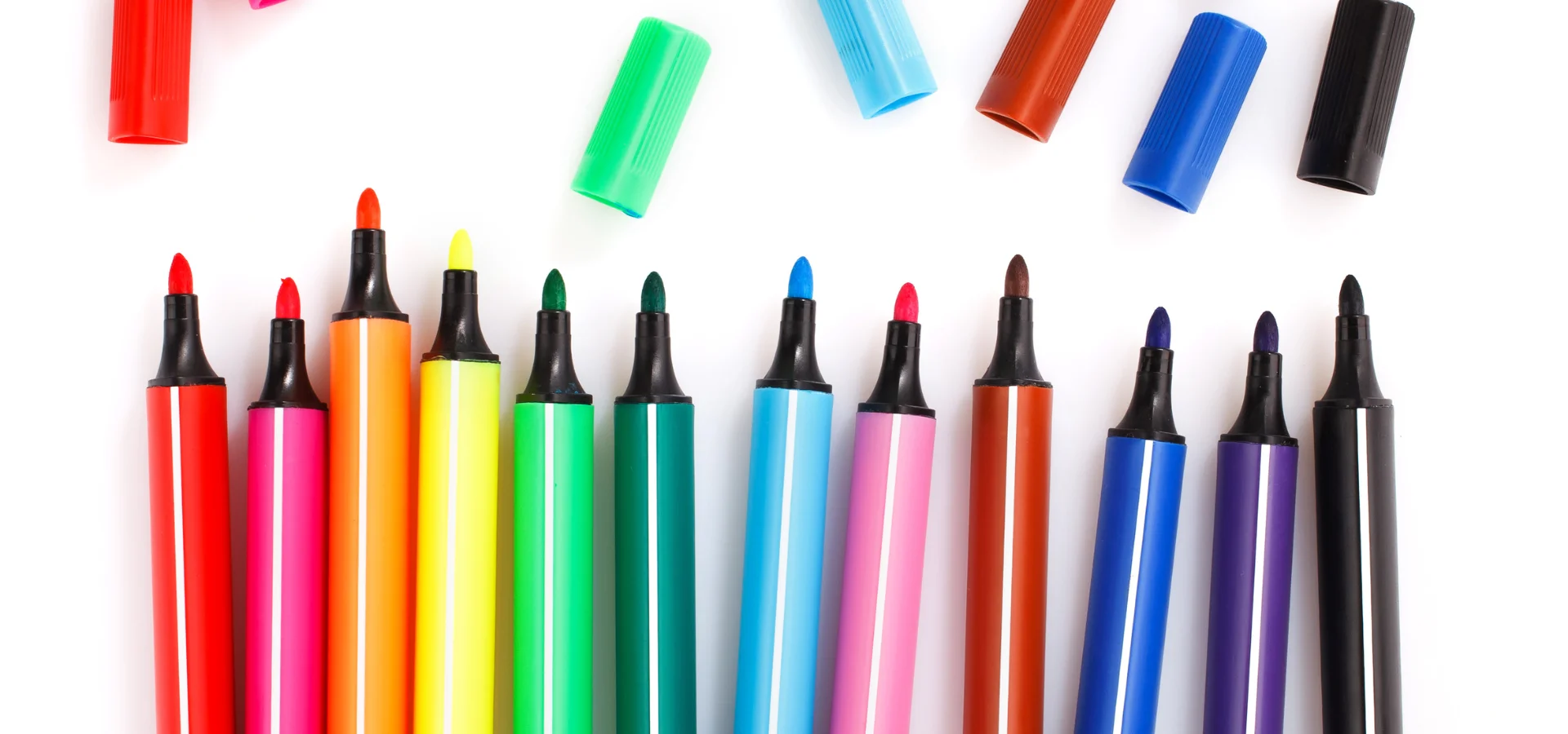 exploring-fabric-markers. in the center of the image there is a line of different coloured markers all facing upwards. they are not neatly in a row. thye are all differnet hieghts. their caps are all off and also haphazardly positioned on the image. on a white background