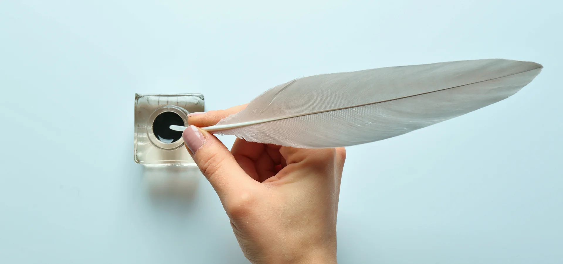 exploring-royal-talens-drawing-ink shows in this image in the centre a hand holding a white quill pen. the feather is white. they are dipping it into a clear glass pot of black calligraphy ink. the light is coming from above them and the shadow is falling under the hand. the background is a baby blue colour