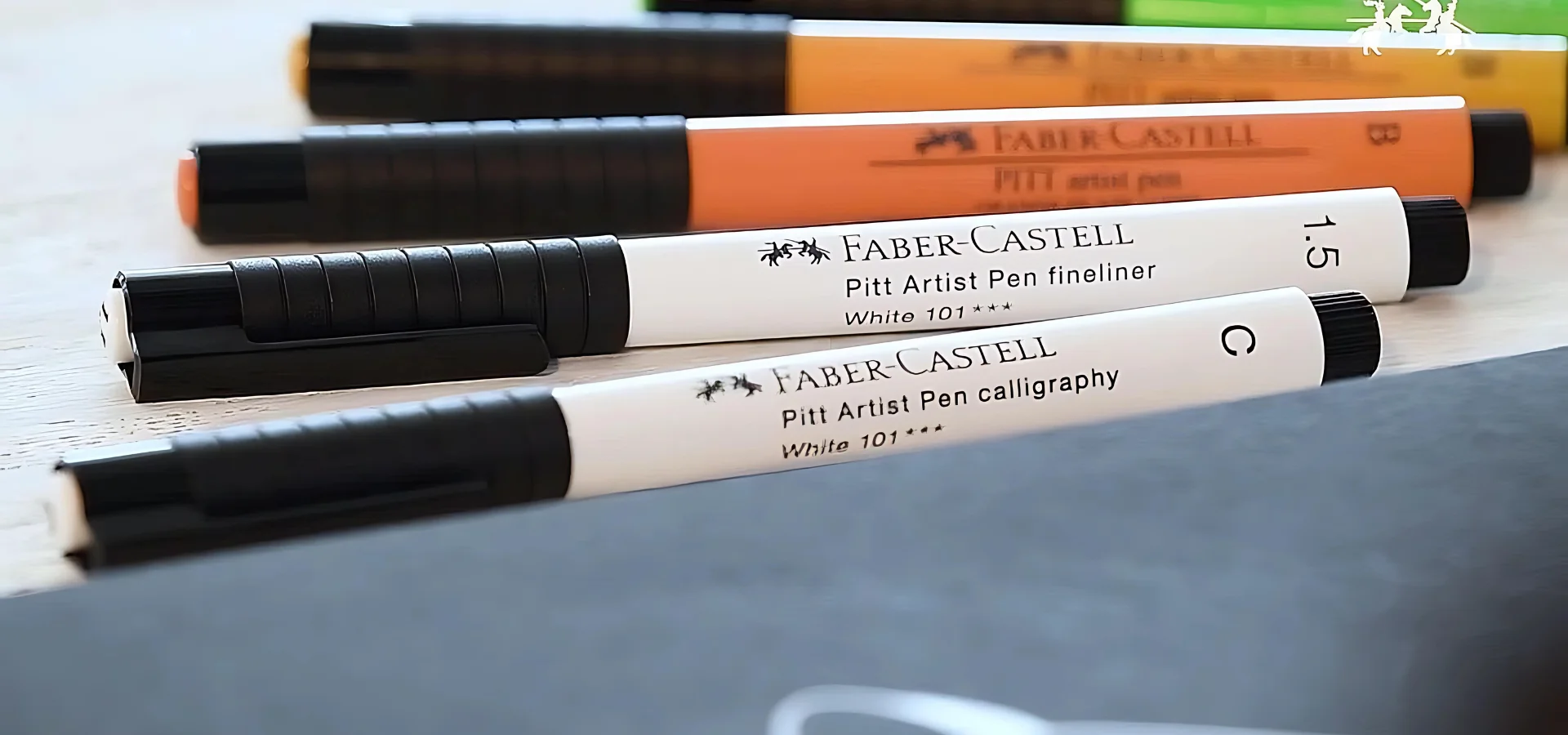 faber-castell-artist-pens-have-you-covered. in the centre of the image there is a book that is closed and just above it lying on their side are five faber castell pens in different colours. they have black lids that are facing towards the left side of the image. they are on a white background