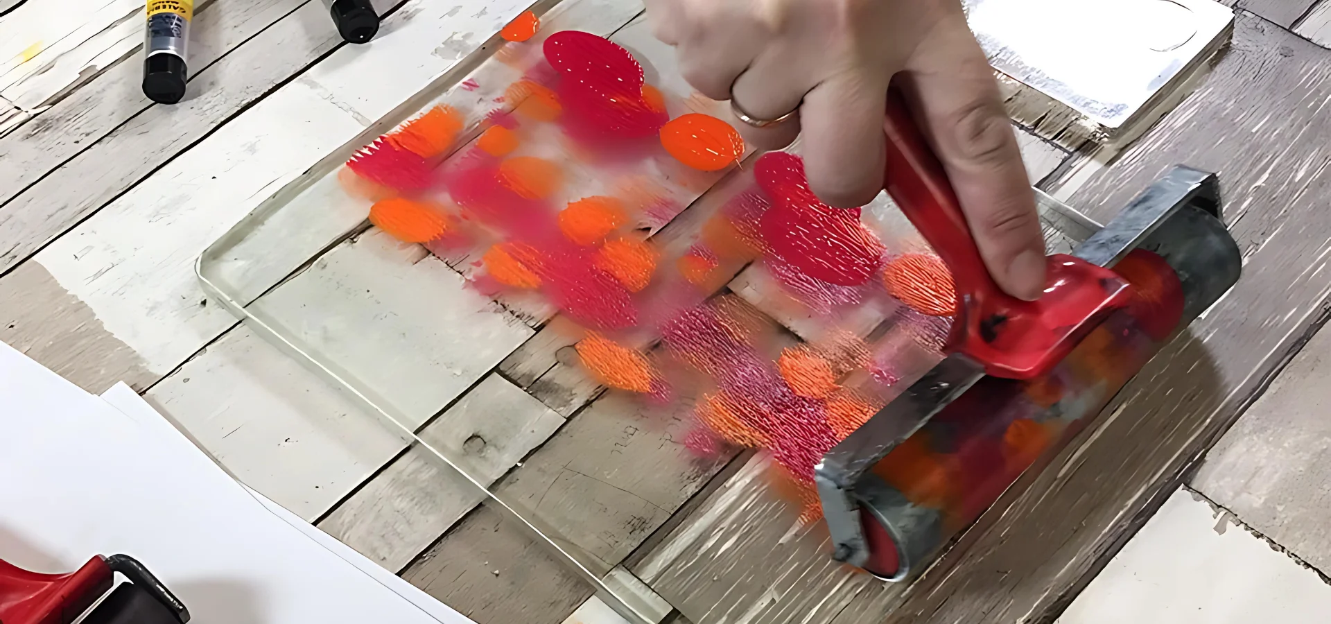 gel-printing-plates-an-artists-secret-weapon. in the center of this image there is a gelli plate that is clear and rectangular and is sitting on a wooden table. there are splotches of pink and orange paint on it and someone is using a brayer to roll the paint over it. on the left hand side on the bottom corner a smaller brayer is visable.