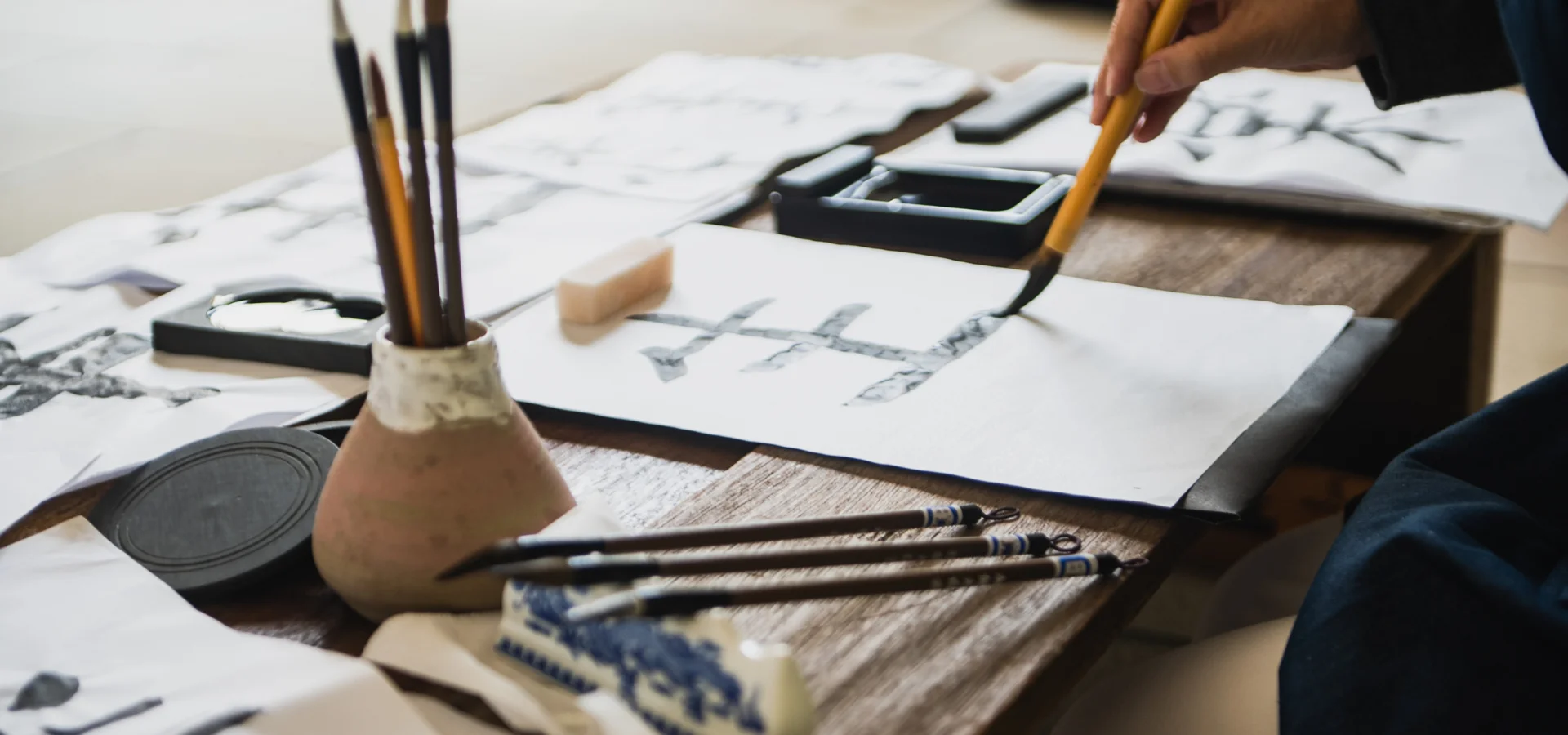 kuretake-sumi-ink-why-you-need-it. in this image there is a person writing in black ink with a paintbrush on a parchment paper on a table that has a pot with used brushes in water and a stand holding other brushes.