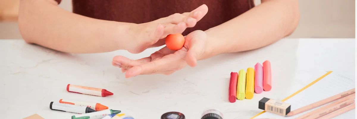 Moulding Marvels Funky Polymer Clay Techniques for Adventure is what this person is doing at the center of the image. there are a pair of hand in the center of the image rolling a blob of orange polymer clay into a ball. on the right hand side of her are four cylindrical shapes that have been rolled out in yellow, orange and red colours. on the left hand side are various shapes that have been cut out of the clay. infront of her are tools being used. she is working on a white table surface