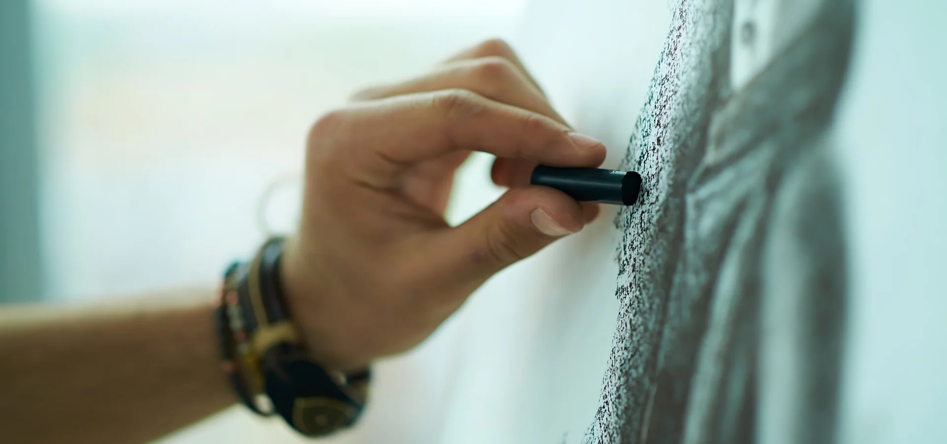 Pitt Monochrome Pastels Unleash your Creativity. in this image you can see a persons hand coming in from the left hand side of the image. they have a bracelt on that is chunky and beaded. they are holding a pastel stick in their hand and they are drawing on a paper. they have already drawn a section as you can see the pastel on the paper. the paper has some shade on it as the light is coming from the opposite end of where the person is standing.