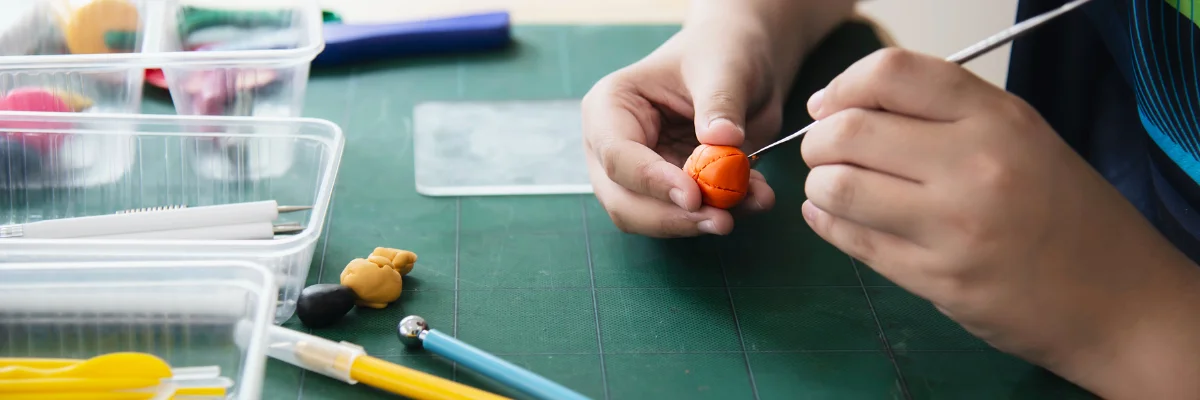 The Claytastic Quest Essential Polymer Clay Tools and Supplies shows a persons hands on the right hand side of the image holding an orange ball of polymer clay in tier left hand and with their right hand using a tool to work on it. on top of a green mat. to the bottom right of the image are some more clay tools and some light orange clay sitting in front of it. the background is a blurred white colour.