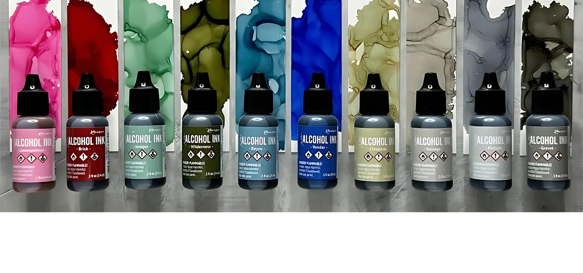 tim-holtz-ranger-alcohol-inks-unveiled. the alcohol inks are lined up against the bottom of the image in a colour order. they are clear bottles with their colour displayed on the label that is wrapped around them. they all have black likds. above them is an ink swtch showing the colour that is inside each bottle. on a wood table background