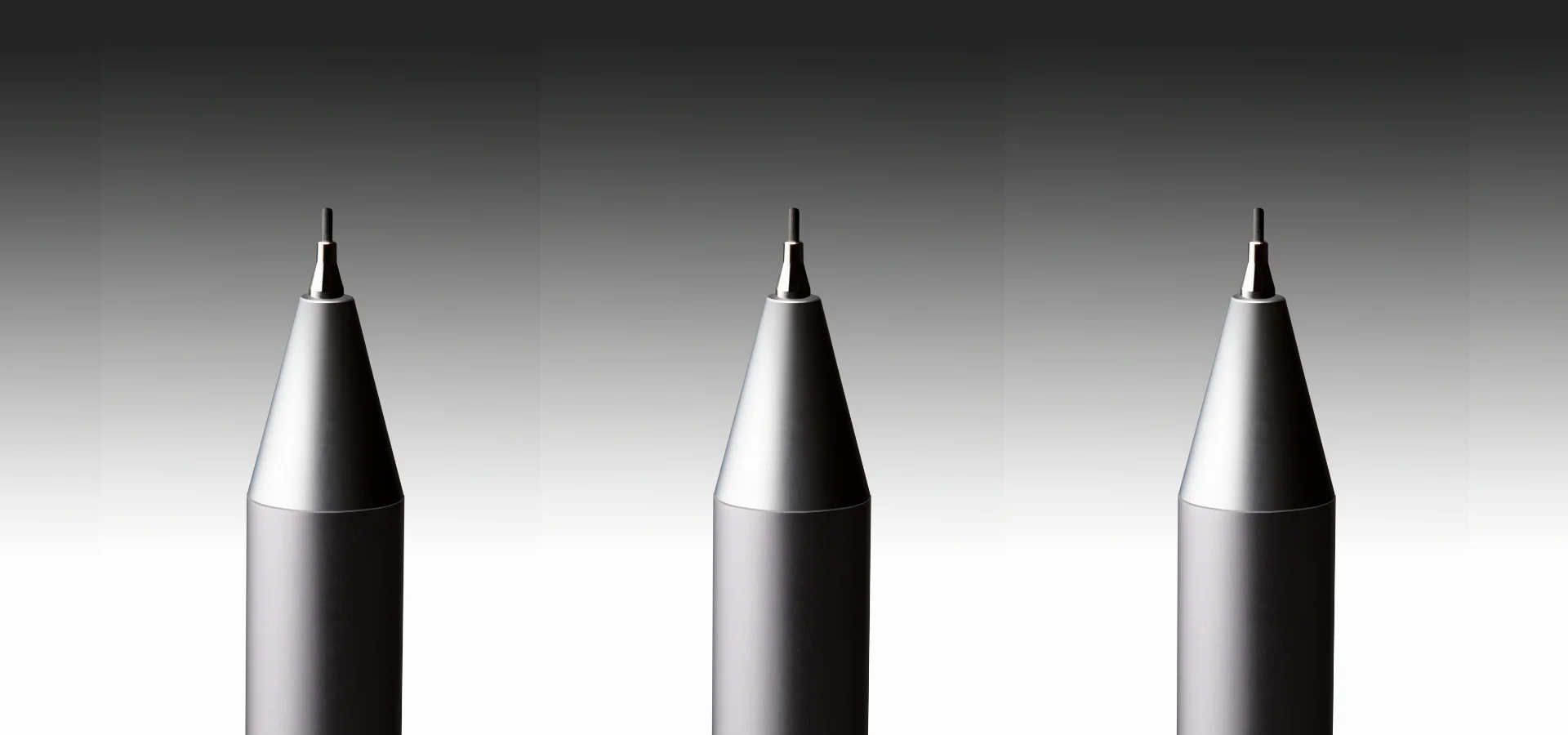 Top-Rated Mechanical Pencils for Drawing Bliss shows three mechanical pencils in a row at the bottom of the image. they are all monotone in black and white. they are tip facing up. it is a dramatic image of clutch pencils. the background is a grey colour going from a darker grey at the top to a light grey towards the bottom