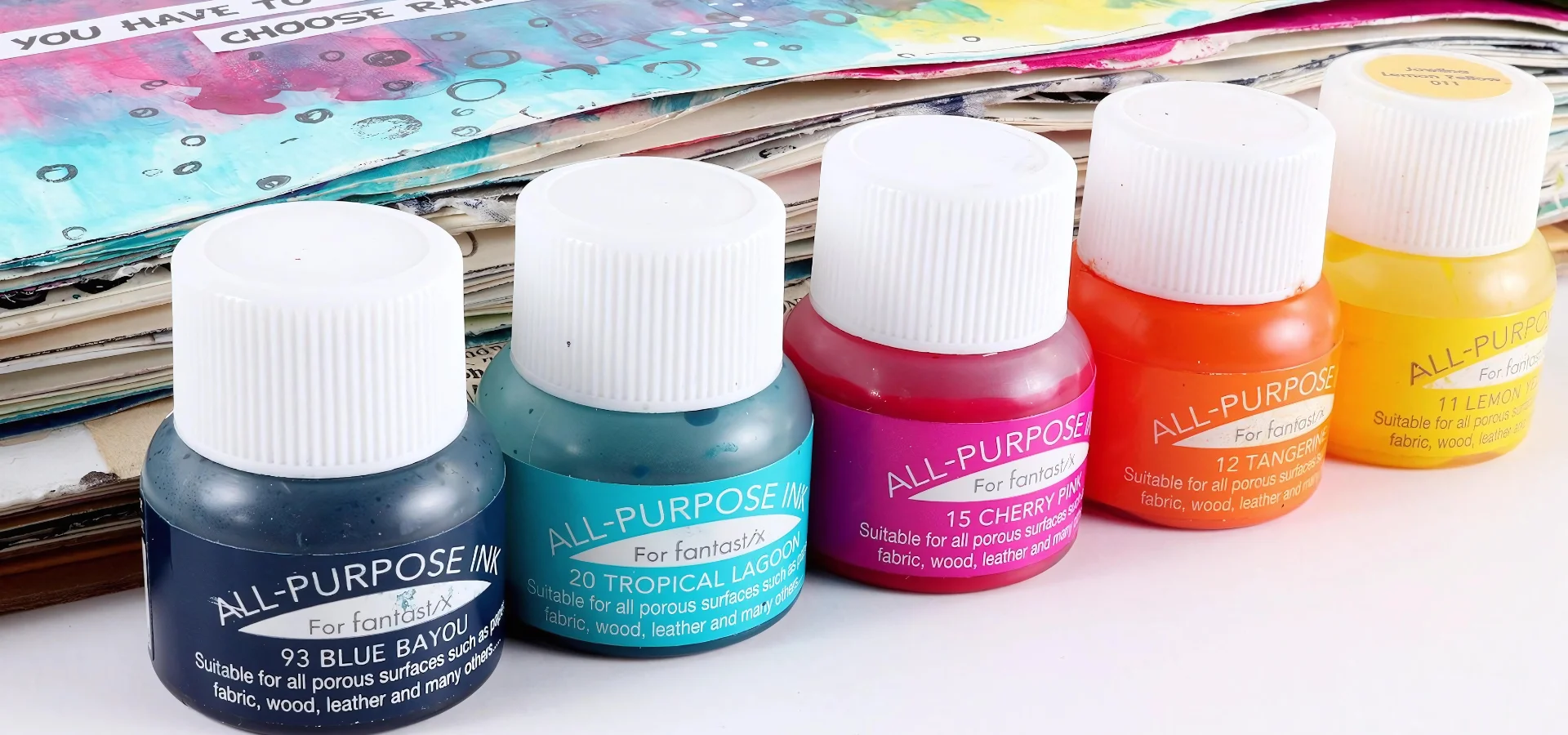 tsukineko-all-purpose-ink-unleash-your-creativity. in this image at the front and running across the image are a row of different coloured bottles of the ink. they are clear bottles showing the colour that is on the inside and their lids are withe. they have labels of the same colour as whats inside with white writing on the,. they are sitting infront of a art journal that has multiple colours on it. on a white table top.