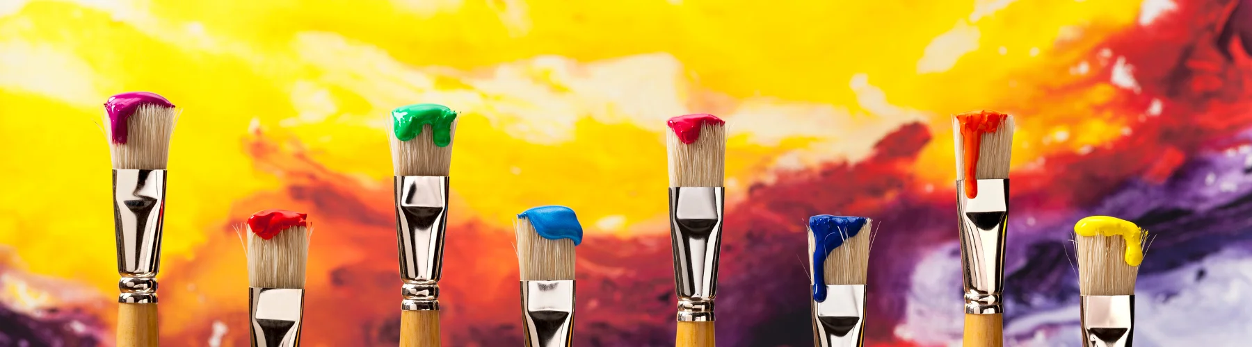 unique-paint-brushes-for-creative-minds. across the bottom of the image are several yellow stemmed brushed with white hairs. all have a blob of different colour paint on them in a rainbow order. they are infront of a rainbow coloured blended background. they have silver ferrules that are shining as the light hits them.