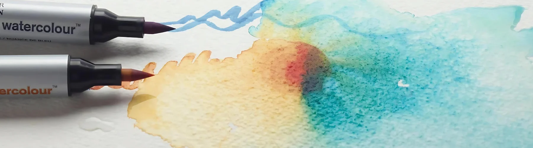 winsor-and-newton-watercolour-markers-deep-dive. in the center of the image are two watercolour markers coming in from the left hand side. they have theor caps off shwoing their brush nibs. they are resting on watercolour paper that has been inked on. there is a mix of blue and orange watercolour ink on the paper that has blended together