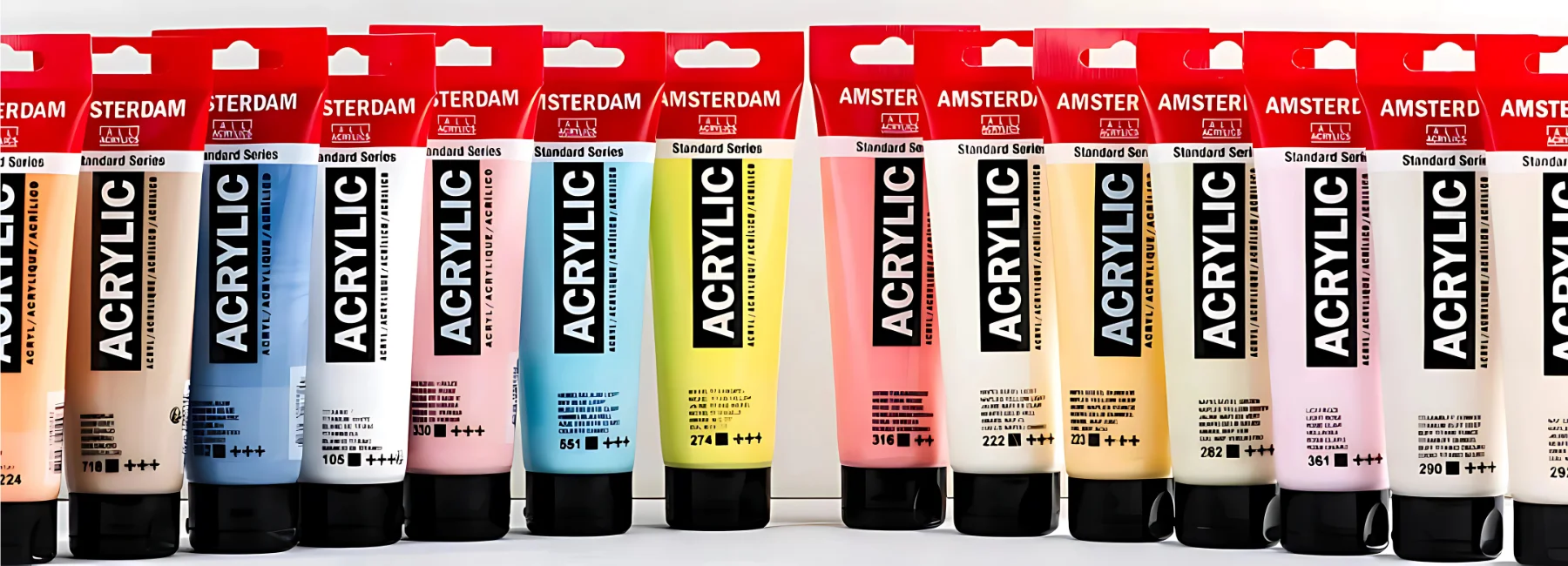 amsterdam-acrylic-paint-beacons-of-artistic-brilliance. across the image there are many tubes of amsterdam acrylic lines up all facing forward in a rainbow order. they all have black caps and are in squeezy tubes. they are clear tubes with a black label in the center and a red top. they are standing on a white table infront of a white background