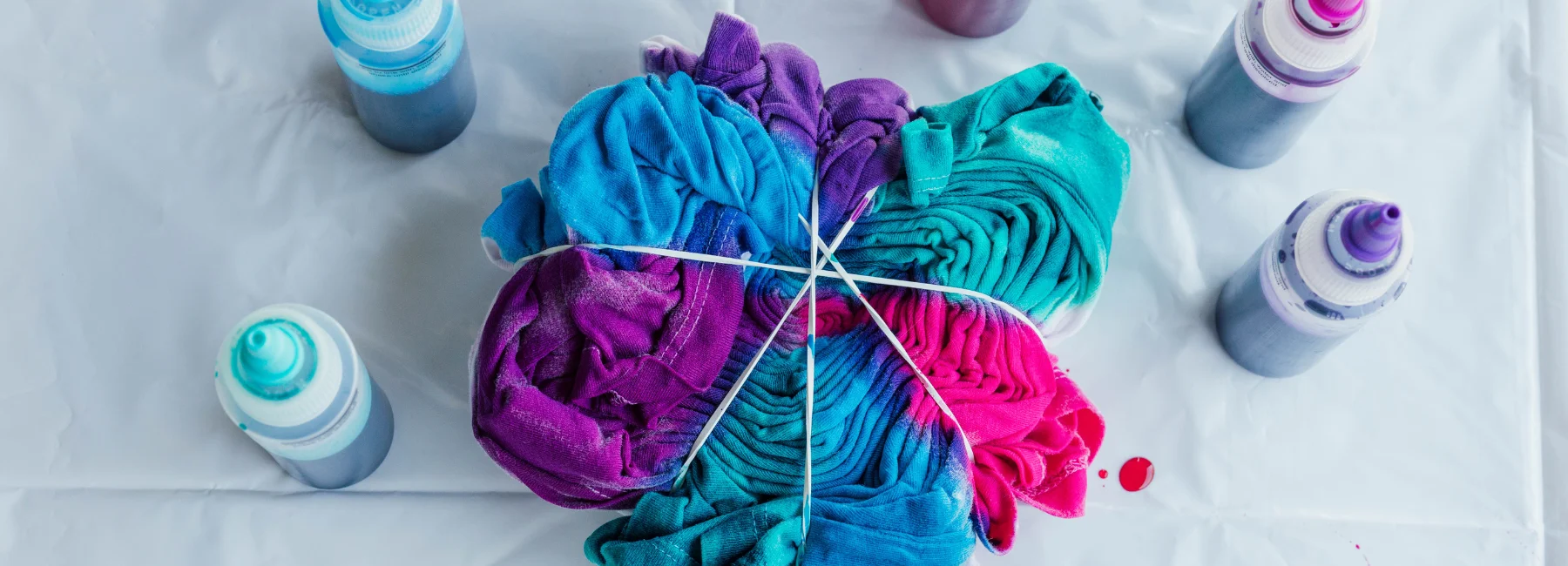fabric-dye-chromatic-adventures. in the center of the image is a shirt that has been bound up by string and elastic bands and has fabric dye poured over it. in the colours of blue, purple and pink. in triangle pattern. around it are tubeds with squeezy lids that have the fabric dye inside of them for application. on a white cloth