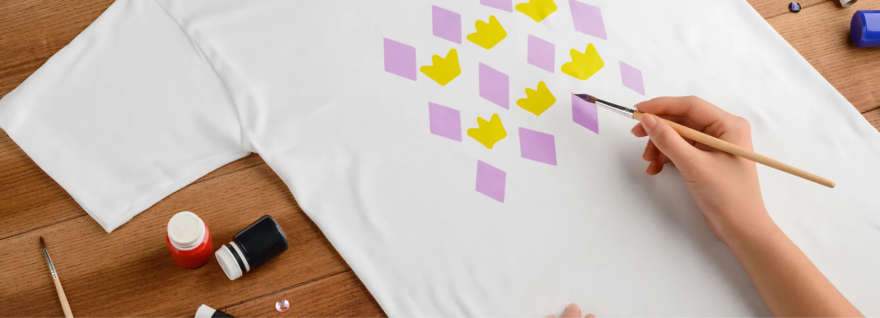 fabric-paint-delights. in the center of the image is a white shirt that is laid out on a table. there is someone holding a paintbrush and painting triangles and crowns onto it. in purple and yellow. with a paintbrush. around them are various bottles of fabric paint scattered on the table with other brushes.