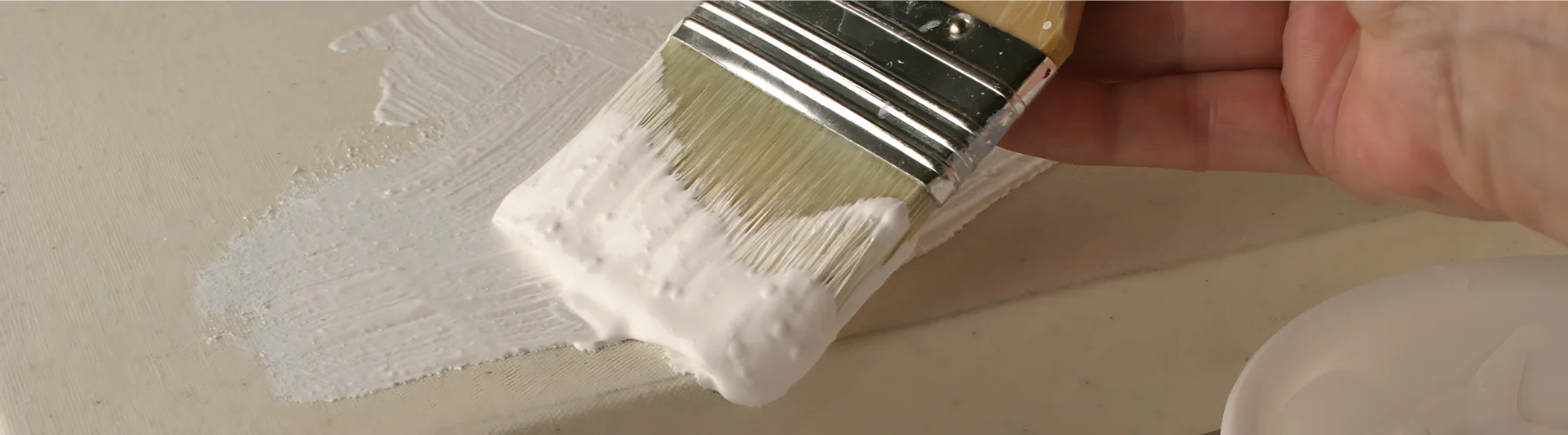 gesso-from-blank-to-swank. the image is a close up of a paintbrush applying the white gesson to a canvas. the paintbrush is full of white gesson and is swiping across the canvas. there is some gesson on the canvs already. a persons hand can be seen on the far right of the image.