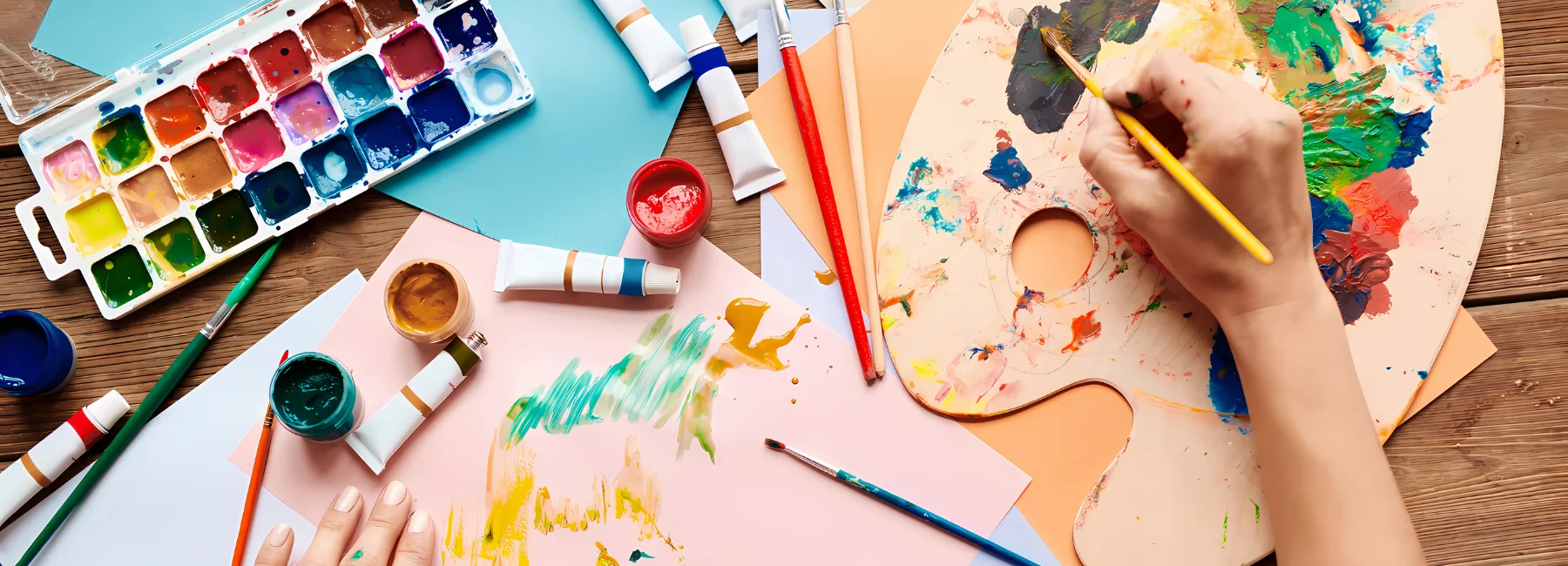 gouache-cakes-painting-paradise-awaits. in the center of the image there is a person mixing gouache colours on a wooden palette that is on the right of the image. on the left is a set of cakes and some tubes scattered around the table. there is paper nad brushes on the table where the person is painting onto.