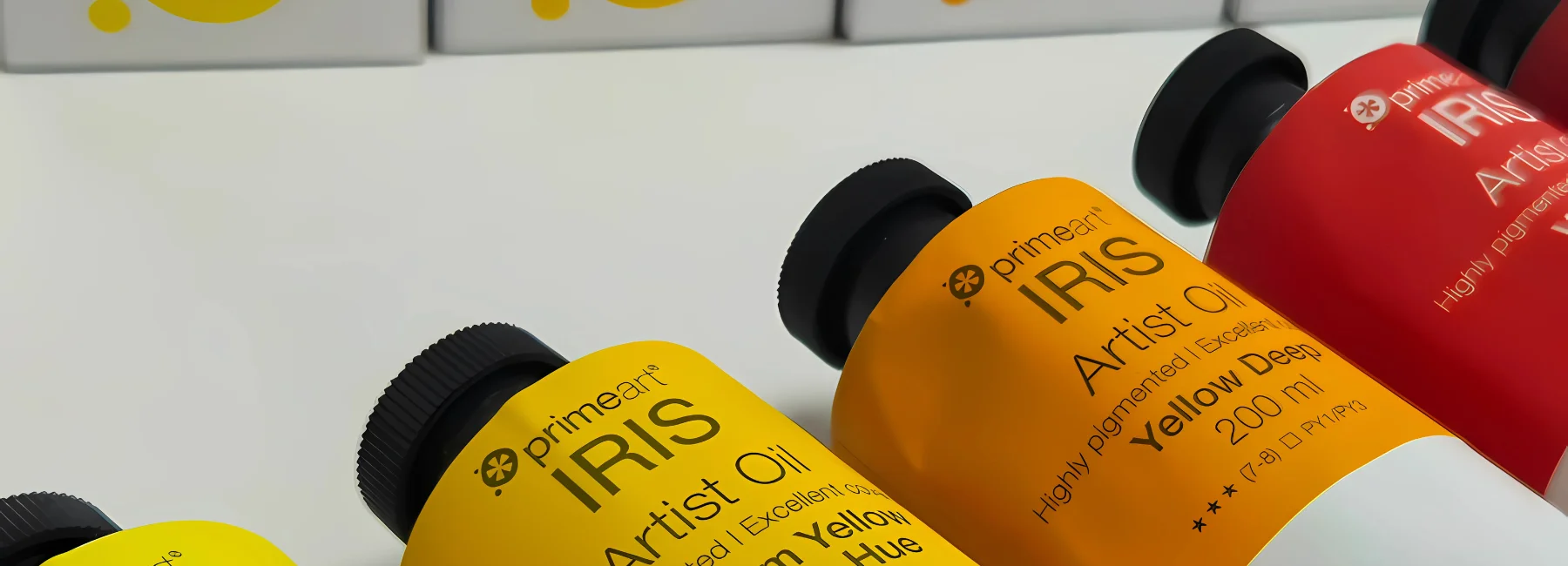 iris-artists-oil-paint-unleash-your-inner-artist. across the centres of the image are the tops of the tubes of iris oil paints. the tops are the colours that are inside the tubes, yellow, orange and red. they all have black caps. behind them are the white boxes that hold the paints. on a white table