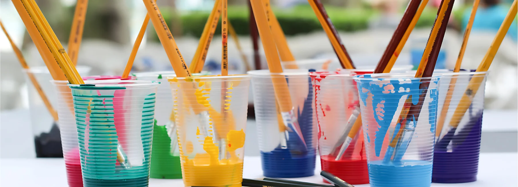 in the center of the image are a collection of clear plastic cups, they all have paint in then filled baout a quarter of the way. there is paint running down the inside of them. the colours are rainbow. there are many paintbrushes sitting hair down inside the cups. the background is a blurred visual of a room.